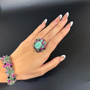 Puede incluir: Una mano muestra un conjunto de anillo y pulsera. El anillo presenta una gran gema ovalada de color verde claro, rodeada de gemas m&aacute;s peque&ntilde;as y coloridas. La pulsera refleja el dise&ntilde;o del anillo, con una disposici&oacute;n similar de piedras preciosas.
