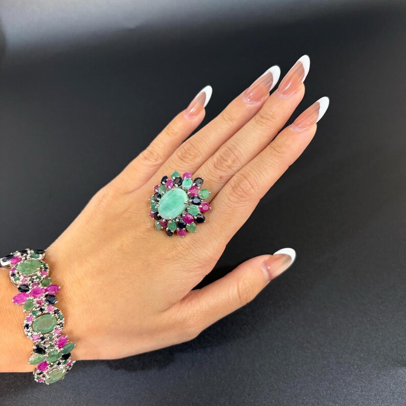 Puede incluir: Una mano muestra un conjunto de anillo y pulsera. El anillo presenta una gran gema ovalada de color verde claro, rodeada de gemas m&aacute;s peque&ntilde;as y coloridas. La pulsera refleja el dise&ntilde;o del anillo, con una disposici&oacute;n similar de piedras preciosas.