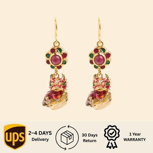 May include: A pair of gold-tone drop earrings featuring a flower-shaped stud with a central red stone surrounded by smaller green and red stones. Below the stud hangs a detailed, multi-colored dragon-like creature in shades of red, green, and gold.
