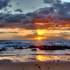 May include: A scenic sunset over the ocean with a dramatic sky of orange, red, and purple clouds. The sun is setting behind the horizon, casting a golden glow on the water. The waves are crashing on the shore, creating a foamy white line.