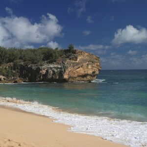 Puede incluir: Una playa de arena con agua turquesa y un acantilado rocoso cubierto de árboles verdes. El cielo es azul con nubes blancas.