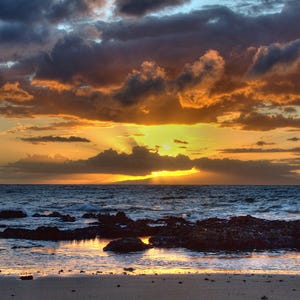 May include: A scenic sunset over the ocean. The sky is filled with orange, yellow, and dark gray clouds. The sun is setting behind the clouds, casting rays of light. The water is dark blue, and there are rocks in the foreground.