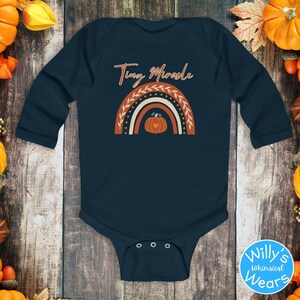 May include: Navy blue baby bodysuit with long sleeves. The bodysuit features a rainbow design with a pumpkin in the center and the words "Tiny Miracle" in a script font. The bodysuit is surrounded by pumpkins and fall foliage.