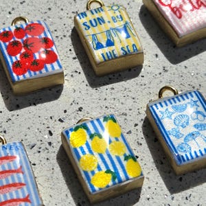 May include: A collection of rectangular pendants with gold-toned metal frames and loops. Designs include red tomatoes on blue stripes, yellow lemons on blue stripes, blue beach items on white, and text reading IN THE SUN BY THE SEA on yellow stripes.