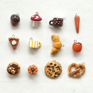 May include: A collection of twelve miniature food and nature charms including an acorn, mushroom, polka dot mug, carrot, slice of pie, striped mug, croissant, tomato, pancake, pumpkin, chocolate chip cookie, and a pretzel, each with a metal loop.