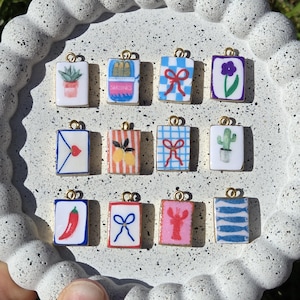 May include: Twelve rectangular metal charms with gold-toned loops, each featuring a unique enamel illustration including a potted plant, a sardine tin, blue bows, a purple flower, an envelope with a heart, lemons, a cactus, a chili, and a lobster.