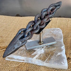 May include: A handcrafted metal sculpture featuring a chain design. The sculpture is made of dark metal and rests on a clear, crystalline base. The chain is held in a metal cradle, creating a unique artistic piece.