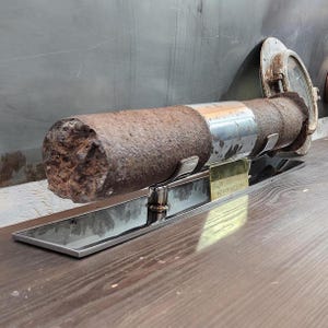 May include: A rusty metal pipe with a silver band is displayed on a black metal stand. The stand has a brass plaque that reads "Intake Gate Pin".