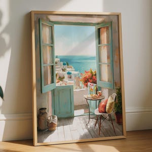 May include: A framed wall art print depicting an open window overlooking a Mediterranean coastal scene with white buildings, blue sea, and a balcony featuring a small round table, a chair with cushions, and vibrant red flowers in a terracotta pot.