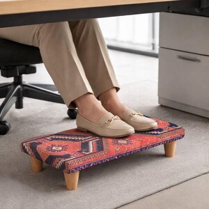 Peut inclure: Repose-pieds rectangulaire orné de motifs rouges, bleus et crème. Il est doté de quatre pieds en bois et est placé sous un bureau. Conçu pour le confort et le soutien pendant le travail.