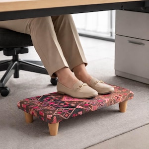 May include: A patterned footrest with wooden legs is positioned under a desk. The footrest features a colorful geometric design in shades of red, pink, and black. The person's feet are resting on the footrest while they are working on a laptop.