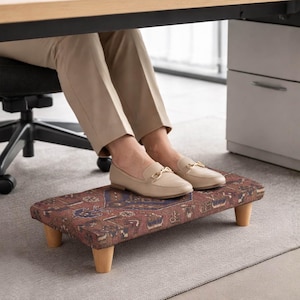 Peut inclure: Un repose-pieds rectangulaire avec un tissu marron-rouge à motifs et quatre pieds en bois. Le repose-pieds est placé sous un bureau, les pieds d'une personne reposant dessus. La personne porte des mocassins beiges.