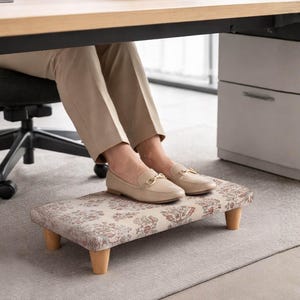 Könnte beinhalten: Ein rechteckiger Fußhocker mit floralem Muster und Holzbeinen unter einem Schreibtisch. Er bietet Unterstützung für die Füße. Die Person trägt beige Loafer. Der Hocker ist etwa 40 cm lang.