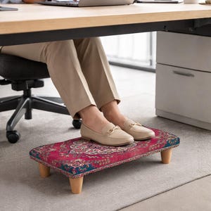 Könnte beinhalten: Ein rechteckiger Fußhocker mit einem roten und blauen Muster, getragen von vier Holzbeinen. Der Hocker steht unter einem Schreibtisch, auf dem die Füße einer Person mit beigen Loafern ruhen.