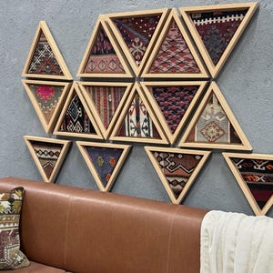 May include: A collection of triangular wall art pieces with wooden frames. Each frame holds a section of patterned fabric in various colors, including red, blue, and beige. The art is mounted on a gray wall above a brown leather sofa.