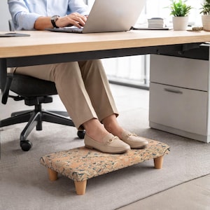 Puede incluir: Un reposapiés rectangular con una parte superior de tela estampada y cuatro patas de madera. El reposapiés está situado debajo de un escritorio, proporcionando apoyo ergonómico. La tela presenta tonos beige, azul y naranja.