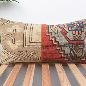 May include: Rectangular decorative pillow with a patterned design. The pillow features a beige and brown geometric pattern on one side and a red, gray, and blue geometric pattern on the other. The pillow is placed on a wooden surface.
