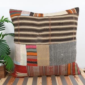 Puede incluir: Un cojín decorativo cuadrado con un diseño de patchwork. El cojín presenta una mezcla de colores y patrones, incluyendo rayas marrones, secciones rojas, naranjas y grises. El cojín está colocado sobre una superficie de listones de madera.