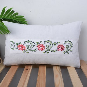 May include: White pillow with a green and pink floral embroidered design. The pillow is on a wooden slatted surface.