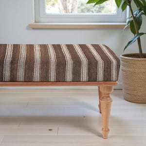 May include: A rectangular bench with a brown and white striped upholstered seat. The bench has four light-coloured wooden legs. A potted plant is visible in the background near a window.