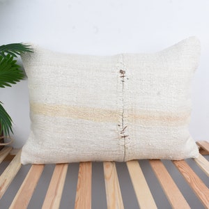 May include: A white throw pillow with a subtle off-white stripe and a raw, unfinished seam down the center. The pillow is made of a soft, textured fabric and is sitting on a wooden slatted surface.