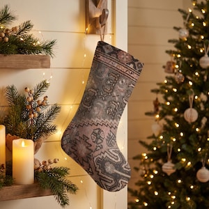May include: A Christmas stocking featuring a muted, multi-colored geometric pattern in shades of dusty pink, charcoal gray, and cream. The stocking has a traditional shape with a hanging loop, displaying a detailed, woven-style textile design.