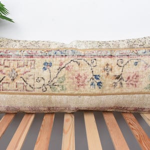 May include: Rectangular beige pillow with a floral pattern in red, blue, and green. The pillow has a woven texture and a decorative border. The pillow is placed on a wooden bed frame.