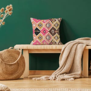 May include: A decorative throw pillow with a geometric pattern in pink, black, gold, and brown tones. The pillow is placed on a wooden bench with a beige blanket draped over it. A woven jute bag and a vase with dried flowers are also in the scene.