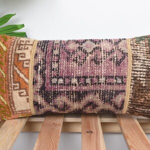 May include: Rectangular decorative pillow with a woven, patterned design. The pillow features a central section in shades of purple and black, flanked by sections in orange and brown. The pillow is placed on a wooden slatted surface.