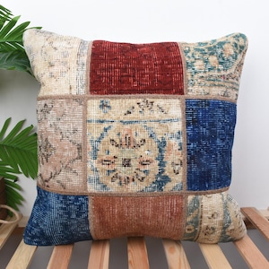 May include: Square patchwork pillow with a variety of colors and patterns. The pillow features a central section of deep red, surrounded by squares of beige, blue, and brown. The pillow is placed on a wooden slatted surface.
