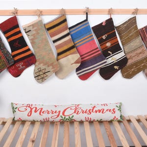 Könnte beinhalten: Acht Weihnachtssocken mit verschiedenen Mustern und Farben, die an einer Holzstange hängen. Ein dekoratives Kissen mit der Aufschrift "Merry Christmas" ist darunter platziert.