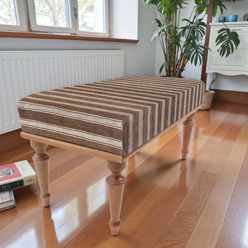 May include: A rectangular bench with a striped brown and beige upholstered seat. The bench has four turned wooden legs. The bench is placed on a hardwood floor. The bench measures approximately 120 cm in length.