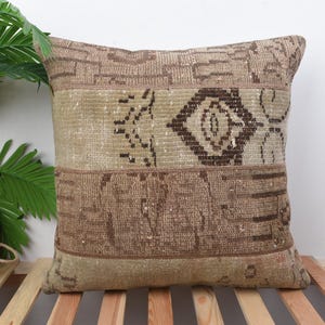 May include: Square decorative pillow with a patchwork design. The pillow features a mix of brown, beige, and tan tones with geometric and abstract patterns. The pillow is placed on a wooden slatted surface.
