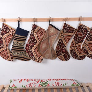 Op de afbeelding: Acht kerstsokken met patronen hangen aan een houten schouw. De sokken zijn gemaakt van bruine stof met rode en gouden accenten. De sokken hebben allemaal verschillende ontwerpen, maar ze hebben allemaal een vergelijkbaar geometrisch patroon.