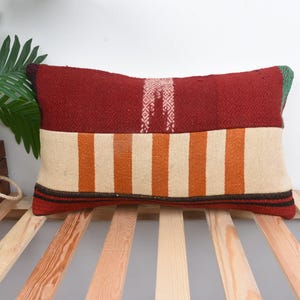 May include: A red, orange, and white striped throw pillow with a green and red border. The pillow is made of woven fabric and has a rustic, handmade look.