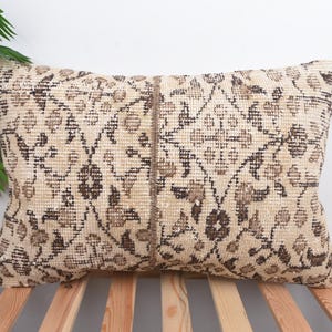 May include: Rectangular beige pillow with a brown floral pattern. The pillow has a vintage, distressed look, with a central vertical seam. The pillow is placed on a wooden slatted surface.
