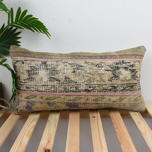 Puede incluir: Una almohada rectangular de color beige con un patrón geométrico rosa y verde. La almohada está hecha de tejido tejido y tiene un borde con flecos.