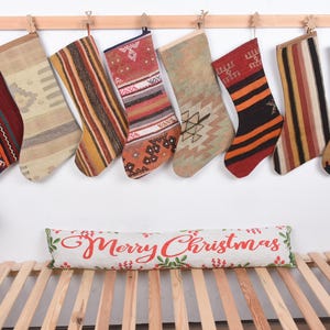 Op de afbeelding: Acht kerstsokken met verschillende patronen en kleuren hangen aan een houten roede. De sokken zijn gemaakt van geweven stof met ontwerpen in rood, bruin, beige en oranje. Een kussen met de tekst "Merry Christmas" ligt eronder.