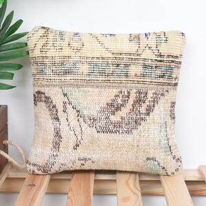 May include: A rectangular, beige pillow with a patterned design. The pillow features a woven texture with brown and blue accents. The pillow is placed on a wooden surface, with a wooden box and greenery in the background.