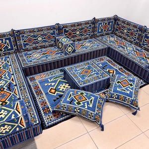 May include: A blue sectional sofa with matching cushions and a rug. The set features geometric patterns in orange, white, and green. The sofa and cushions have a striped border. The rug and cushions have tassels.
