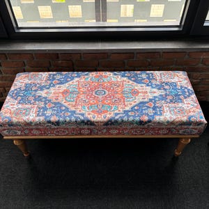 Könnte beinhalten: Eine dekorative Bank mit einer bunten, gemusterten Stoffoberfläche. Der Stoff zeigt ein blaues, rotes und oranges geometrisches Muster. Die Bank hat Holzbeine.