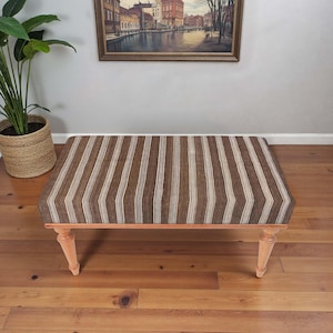May include: A rectangular ottoman with a brown and cream striped fabric top. The ottoman has four light-coloured wooden legs and a wooden frame. The piece is placed on a hardwood floor.