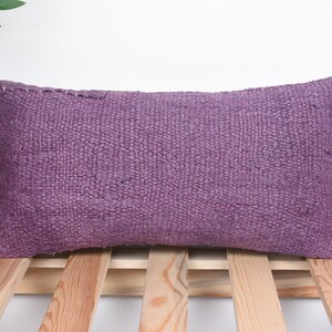 May include: A rectangular, purple woven pillow with a textured surface. The pillow is placed on a wooden slatted surface. The pillow is a solid color and appears to be made of a natural fiber.
