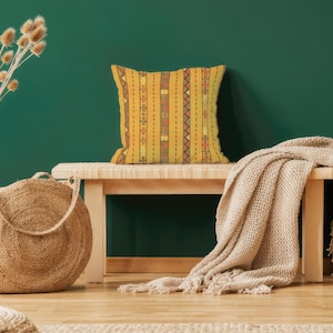 May include: A decorative yellow pillow with vertical striped patterns and geometric designs rests on a wooden bench. A woven beige blanket is draped over the bench. A large woven tote bag and a glass vase with dried plants are in the foreground.