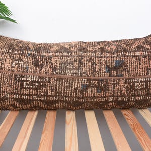 May include: A rectangular, brown and beige patterned pillow with a textured appearance. The pillow has a dark brown band across the center. The pillow is placed on a wooden bed frame.