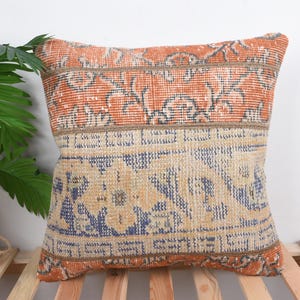 May include: Square cushion with a vintage rug design. The cushion features orange, beige, and blue colours. The top and bottom sections have an orange pattern, while the centre has a beige and blue pattern.
