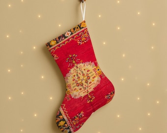 Puede incluir: Un calcetín navideño rojo con un diseño estampado, colgado de un gancho de madera. El calcetín presenta un motivo floral central en amarillo y blanco, con bordes negros, amarillos y rojos. Se utiliza un lazo de cinta blanca para colgarlo.