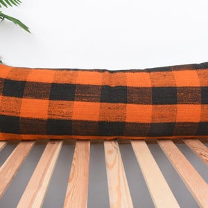 May include: An orange and black plaid lumbar pillow. The rectangular pillow features a bold buffalo check pattern. The pillow is placed on a wooden bed frame, with a white background and a green plant in the upper left corner.