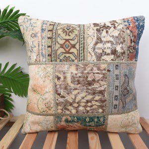 Puede incluir: Cojín cuadrado de patchwork con diseño de alfombra vintage. El cojín presenta una mezcla de patrones beige, marrón, azul y óxido. El cojín está colocado sobre una superficie de listones de madera, con una planta en maceta en el fondo.