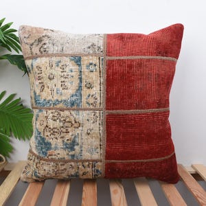 May include: Square cushion with a patchwork design. One half has a vintage rug pattern in beige, blue, and brown. The other half is a solid, textured, red fabric. The cushion is placed on a wooden slatted surface.
